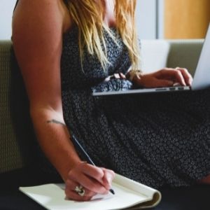 Eine Frau sitzt auf der Couch mit dem Laptop und schreibt etwas parallel auf