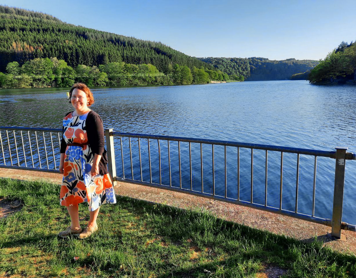 Yvonne steht auf einer Wiese vor einem See und trägt ein Blumenkleid